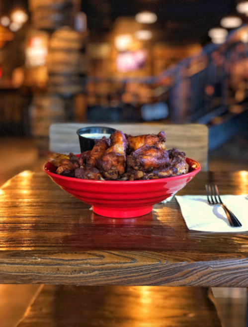A red bowl filled with crispy chicken wings sits on a wooden table, with a small cup of sauce nearby.