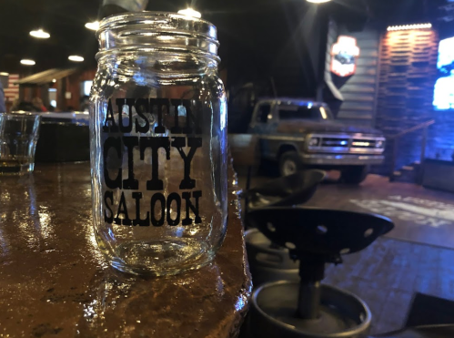A glass jar labeled "Austin City Saloon" sits on a bar, with a vintage truck and dimly lit background.