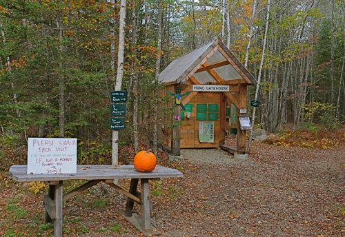 A wooden gatehouse in a forest with a pumpkin on a bench and a sign asking visitors to sign in.