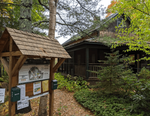 A wooden signpost with maps stands in front of a rustic building surrounded by trees and autumn foliage.