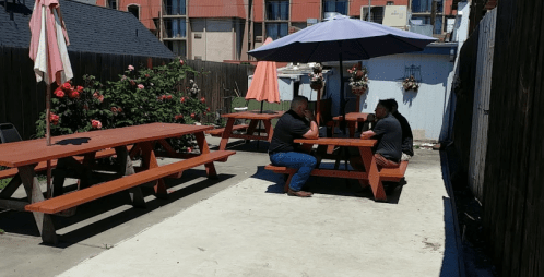 Two people sit at a picnic table under an umbrella in a garden area with flowers and a fence.