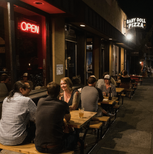 A lively outdoor dining scene at night, with people enjoying pizza at Baby Doll Pizza under an "Open" sign.
