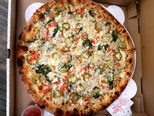 A large pizza topped with spinach, tomatoes, jalapeños, and melted cheese in a pizza box, with a side of dipping sauce.