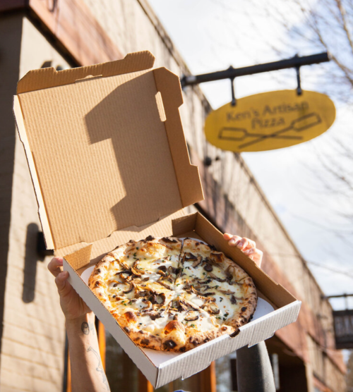 A person holds a pizza box open, revealing a pizza topped with cheese and mushrooms, outside a pizzeria.
