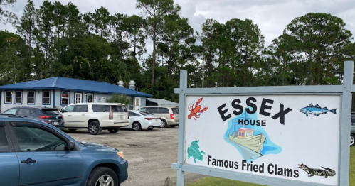 A sign for Essex House, featuring "Famous Fried Clams," with a blue building and parked cars in a wooded area.
