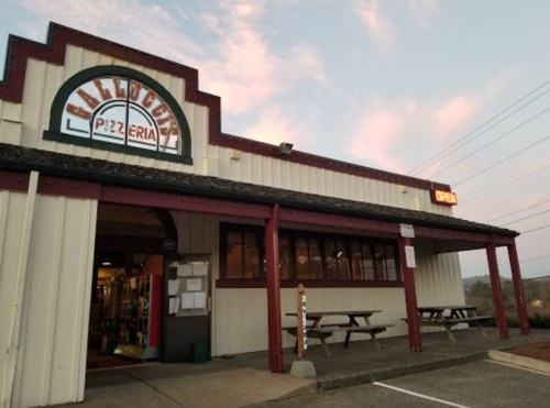 Exterior of a pizzeria with a sign reading "Open," featuring outdoor seating and a colorful sky at dusk.