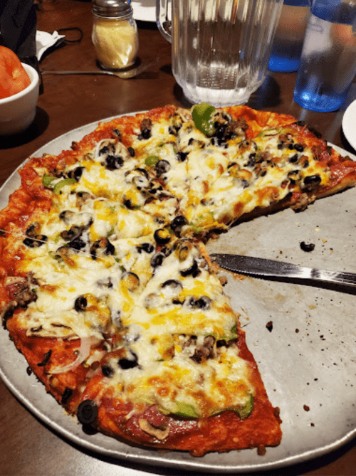 A pizza on a metal tray, topped with cheese, olives, green peppers, and onions, with a few slices removed.