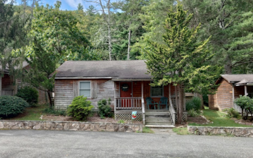 A rustic wooden cabin surrounded by trees, featuring a small porch with two chairs and a stone pathway.