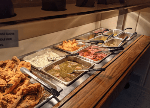 A buffet spread featuring fried chicken, rice, greens, shrimp, and various sides in metal trays on a wooden counter.