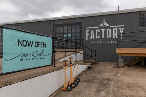Exterior of a building with a sign reading "Now Open Wear It Wool" and "Factory at Columbia," featuring stairs and wooden carts.
