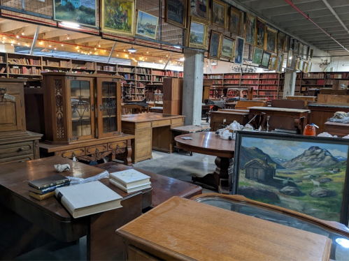 A spacious room filled with wooden furniture and shelves of books, adorned with various paintings on the walls.