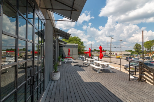 Outdoor patio with picnic tables and red umbrellas, featuring a modern building and a clear blue sky with clouds.