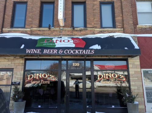 Exterior of Dino's Gourmet Pizzeria, featuring a sign for wine, beer, and cocktails, with snow on the ground.