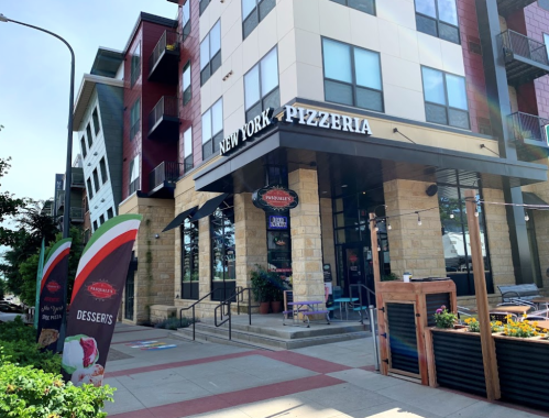 Exterior of a pizzeria named "New York Pizzeria" with colorful banners and a modern building in the background.