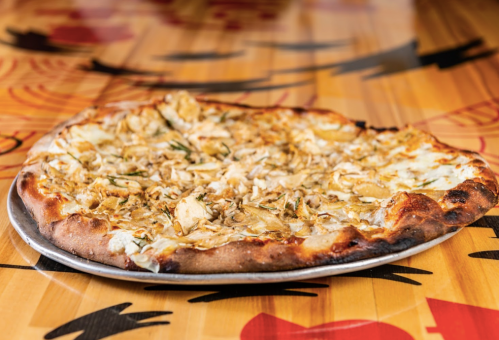 A freshly baked pizza topped with chicken and herbs, served on a silver platter against a colorful wooden table.