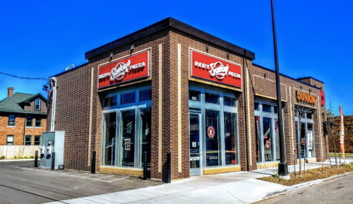 Exterior of a brick building featuring a pizza restaurant with large windows and a bright red sign. Clear blue sky above.