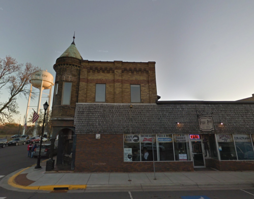 Historic brick building with a turret, water tower in the background, and storefronts along a quiet street at dusk.