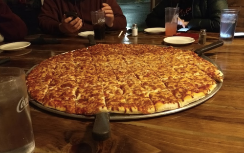 A large, round pizza with melted cheese and sliced into pieces, surrounded by drinks and people at a table.