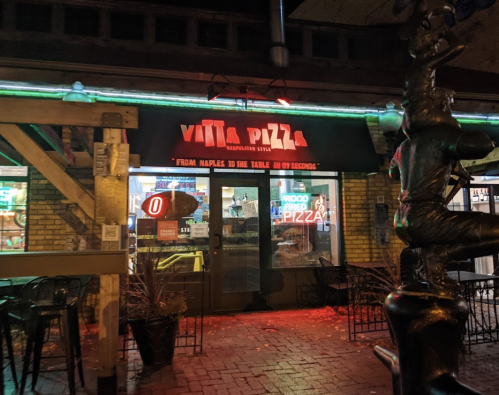 A pizza restaurant entrance with neon lights and a sign reading "Vita Pizza" in a lively outdoor setting.