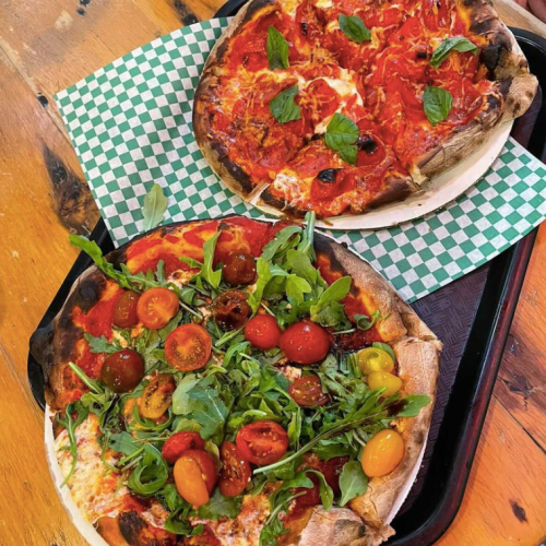Two pizzas on a tray: one topped with tomato sauce and basil, the other with cherry tomatoes and arugula on a green checkered paper.