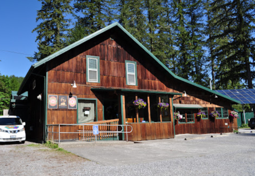A rustic wooden building surrounded by trees, featuring large windows and flower baskets, with a gravel parking area.