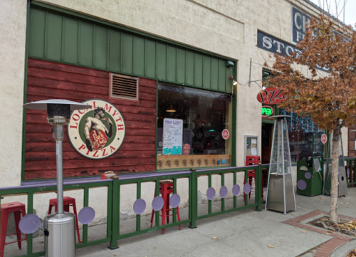 Exterior of Local Myth Pizza with a green awning, outdoor seating, and a sign on the wall.