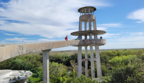A concrete observation tower with a walkway, surrounded by lush greenery and a clear blue sky.