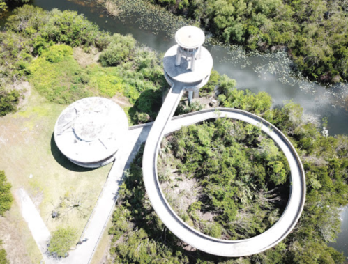 Aerial view of a circular walkway leading to a tower, surrounded by lush greenery and a winding waterway.