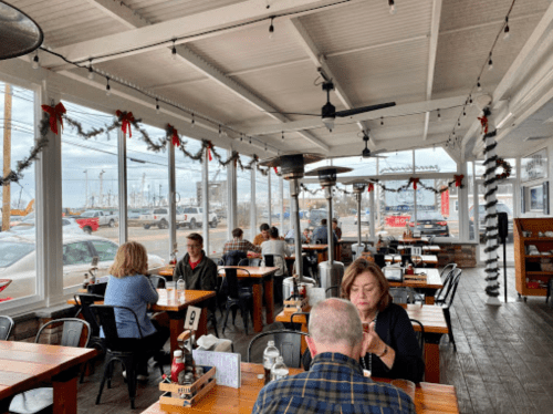 A cozy restaurant with wooden tables, patrons dining, and festive decorations, overlooking a harbor view.