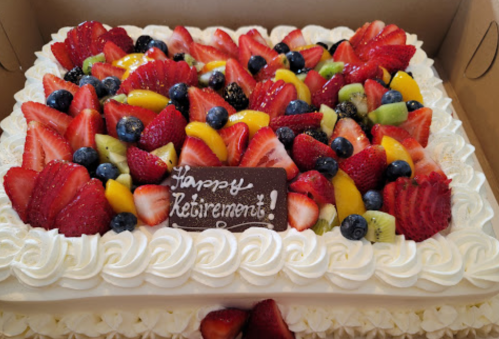 A decorated retirement cake topped with fresh strawberries, blueberries, kiwi, and a chocolate plaque that says "Happy Retirement!"