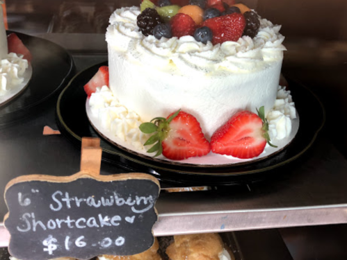 A 6-inch strawberry shortcake topped with fresh berries and whipped cream, displayed in a bakery case. Price: $16.00.