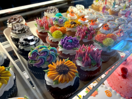 A display of colorful cupcakes with intricate floral frosting designs in a bakery case.