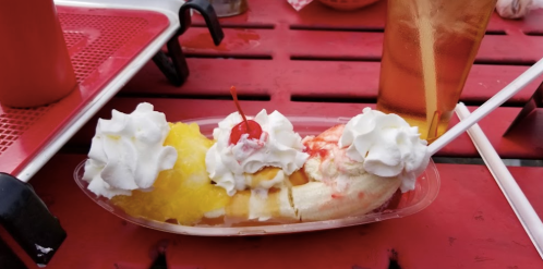 A colorful banana split topped with whipped cream, cherries, and pineapple, served on a red table.