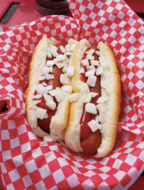 Two hot dogs in a bun topped with ketchup and chopped onions, served in a red and white checkered paper basket.