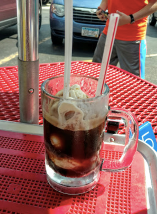 A frosty mug of root beer float with two straws, sitting on a red table in a casual outdoor setting.