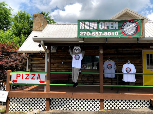 A log cabin pizza shop with a raccoon mascot and T-shirts displayed, featuring a "Now Open" sign.