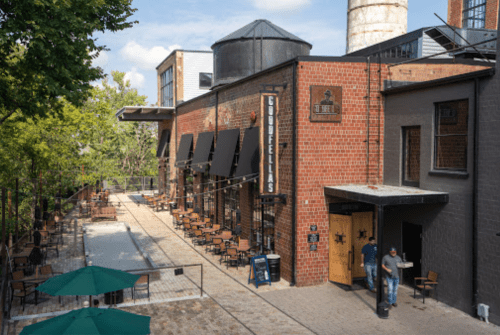 A brick building with outdoor seating, shaded by umbrellas, and a person entering through a door. Trees in the background.