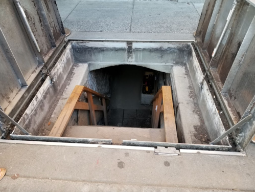 A view of stairs leading down into a dimly lit underground entrance. The surrounding area is concrete and weathered.