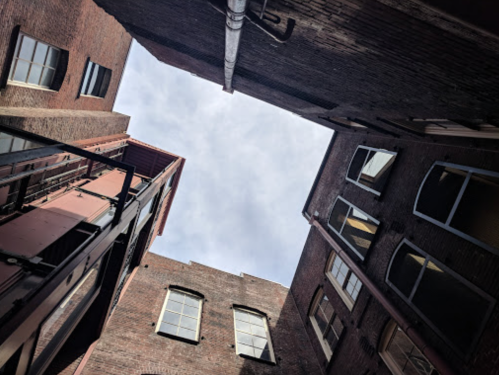 View looking up between brick buildings, with a cloudy sky visible above.