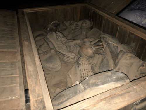 A wooden box filled with various old, dusty boots and shoes, partially covered in dirt.