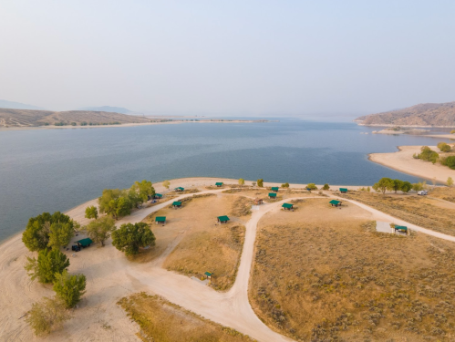 Aerial view of a serene lakeside campsite with green tents, sandy paths, and surrounding hills under a hazy sky.