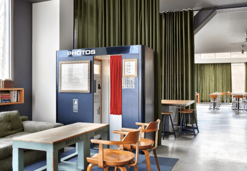 A cozy interior featuring a photo booth, green curtains, wooden chairs, and a rustic table in a modern space.
