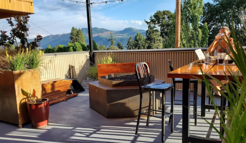 A scenic outdoor dining area with a fire pit, table, and a person enjoying a drink against a mountain backdrop.