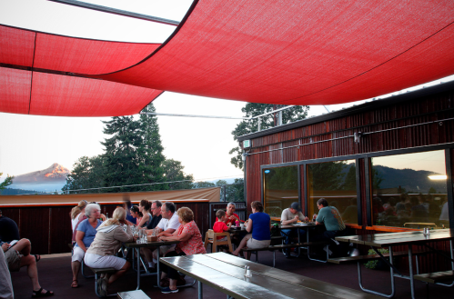 A lively outdoor dining area with people gathered at tables, shaded by red canopies, with mountains in the background.