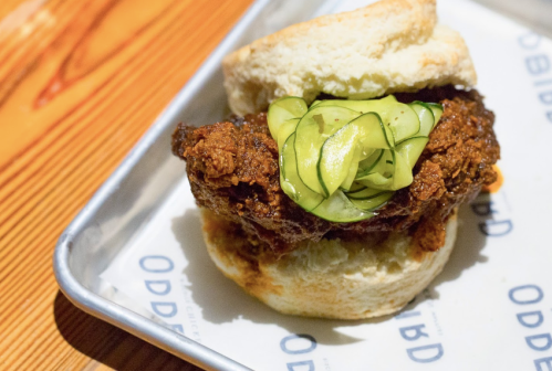 A crispy fried chicken sandwich with pickles on a soft biscuit, served on a metal tray.