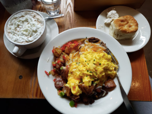 A hearty breakfast plate with scrambled eggs, potatoes, peppers, and a biscuit, alongside a cup of whipped cream-topped coffee.