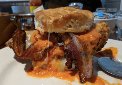 A hearty biscuit sandwich filled with fried chicken, crispy bacon, and drizzled with a spicy sauce.