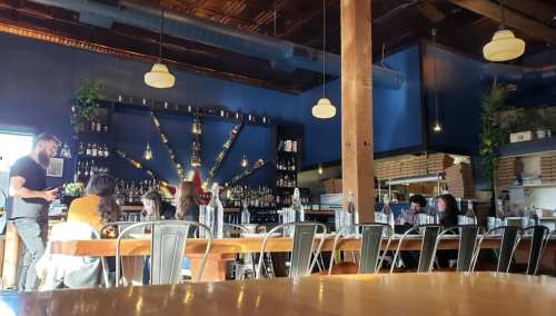 A cozy restaurant interior with wooden tables, shelves of bottles, and patrons enjoying their meals.