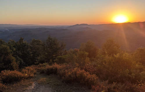 A serene mountain landscape at sunset, with rolling hills and trees silhouetted against a colorful sky.