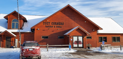 A wooden building with a sign reading "Cozy Corners Tavern & Grill," surrounded by snow and a parked car.